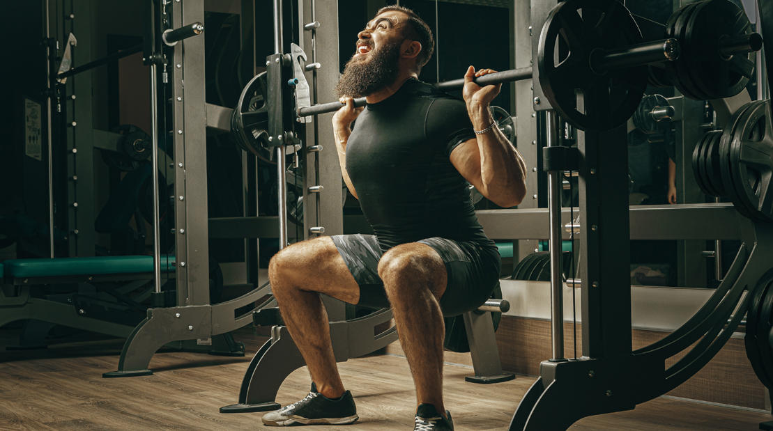 Bearded weight lifter struggling with combination exercises with a squat at smith machine workout and performing squat accessory exercises